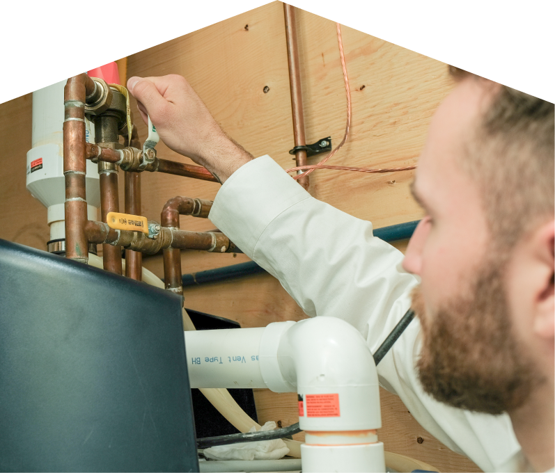 Romaniuk water heater specialist tightens a valve on copper piping during a water heater installation, with exposed wood paneling and wiring in the background.
